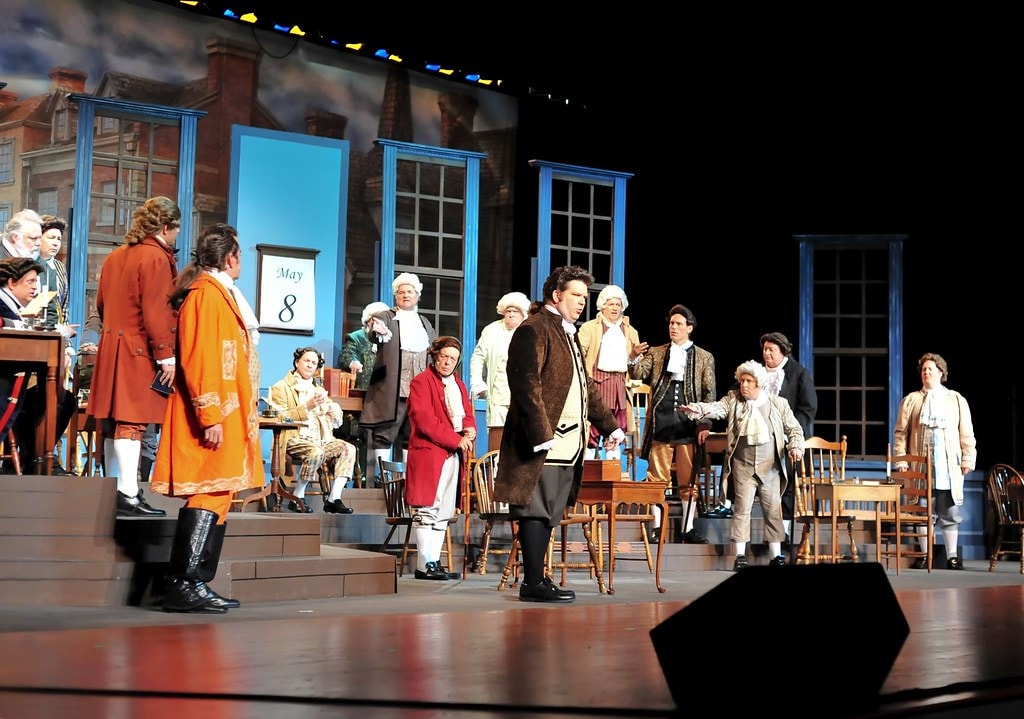 1776 — the full ensemble of delegates in 18th-century colonial coats and wigs fills the Continental Congress chamber set, a May 8 calendar visible on the wall, as a delegate addresses the floor