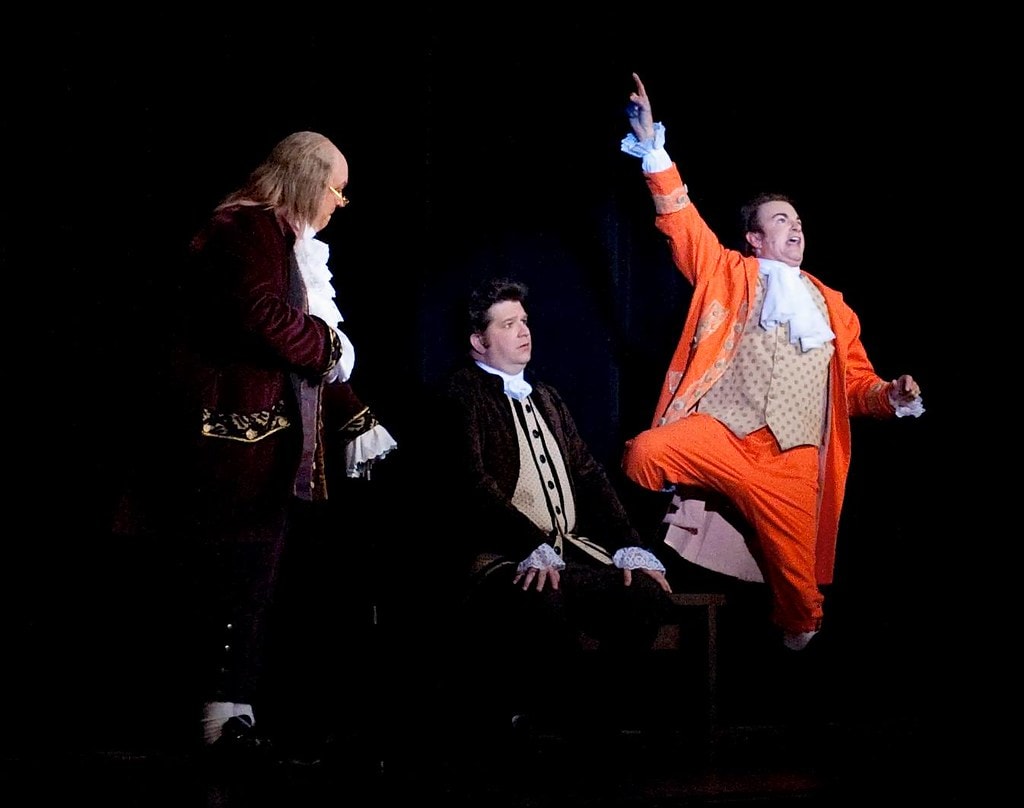 1776 — three men in colonial costume against a black backdrop: a man in a burgundy coat at left, a seated man in dark velvet at center, and a man in a vivid orange suit leaping exuberantly with one arm raised
