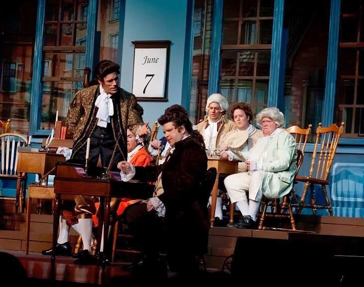 1776 — a delegate in a dark grey coat stands reading a document aloud at a writing desk while another man reclines in a chair listening in the intimate chamber scene