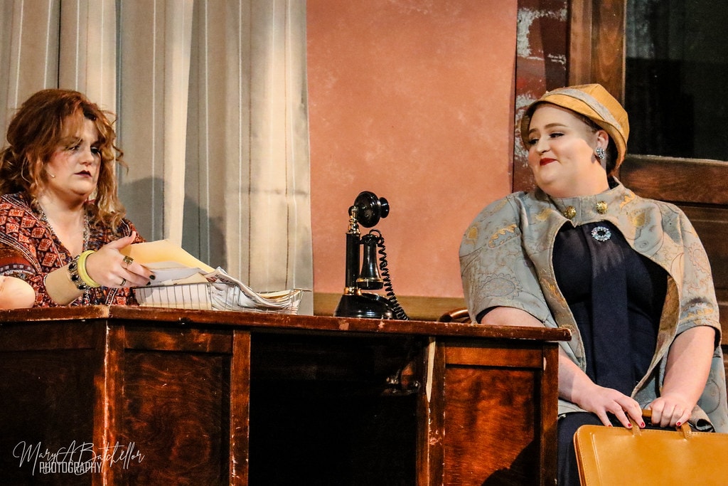 Annie — Miss Hannigan at her wooden desk faces a woman in a teal cape and gold hat