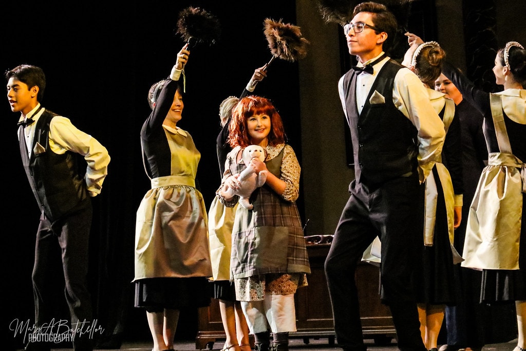 Annie — Annie holds her rag doll and beams as Warbucks mansion staff raise feather dusters