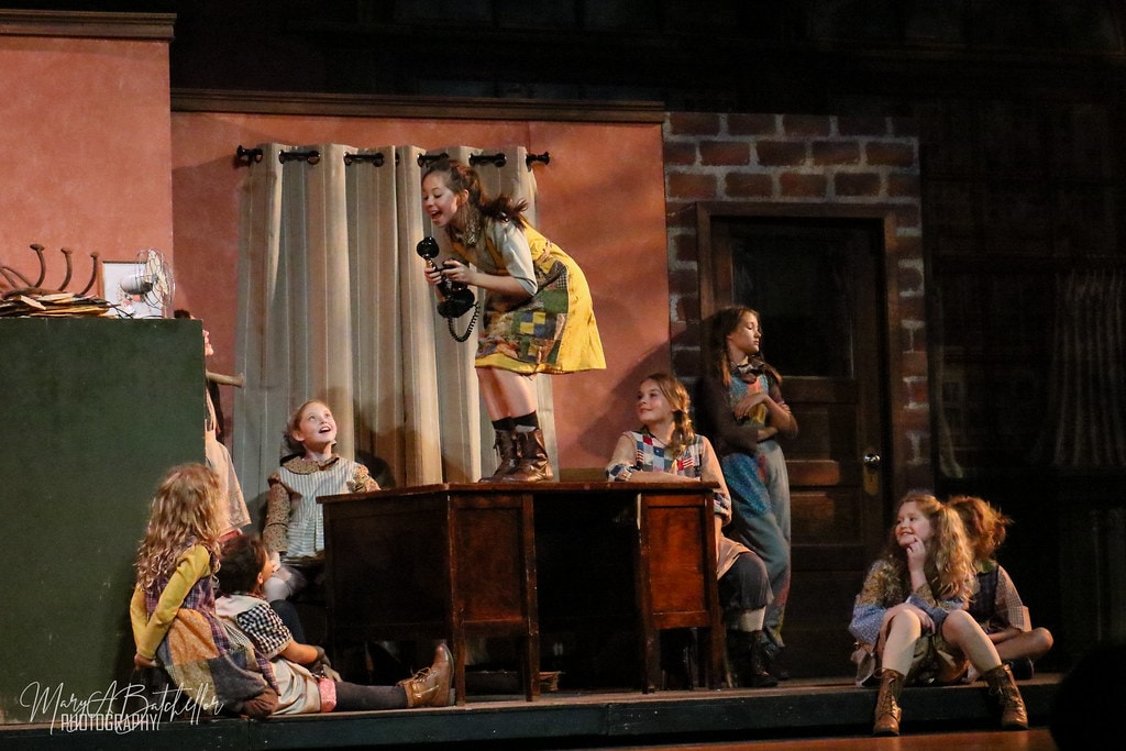 Annie — Annie stands on a wooden cabinet holding a vintage telephone as orphan girls watch