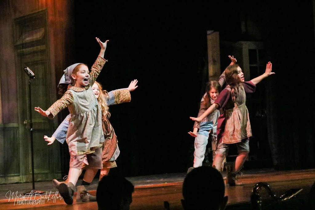 Annie — four orphan girls leap and dance with arms wide before the orphanage backdrop