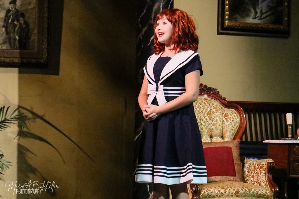 Annie — Annie in a navy sailor dress sings with hands clasped in the Warbucks parlor