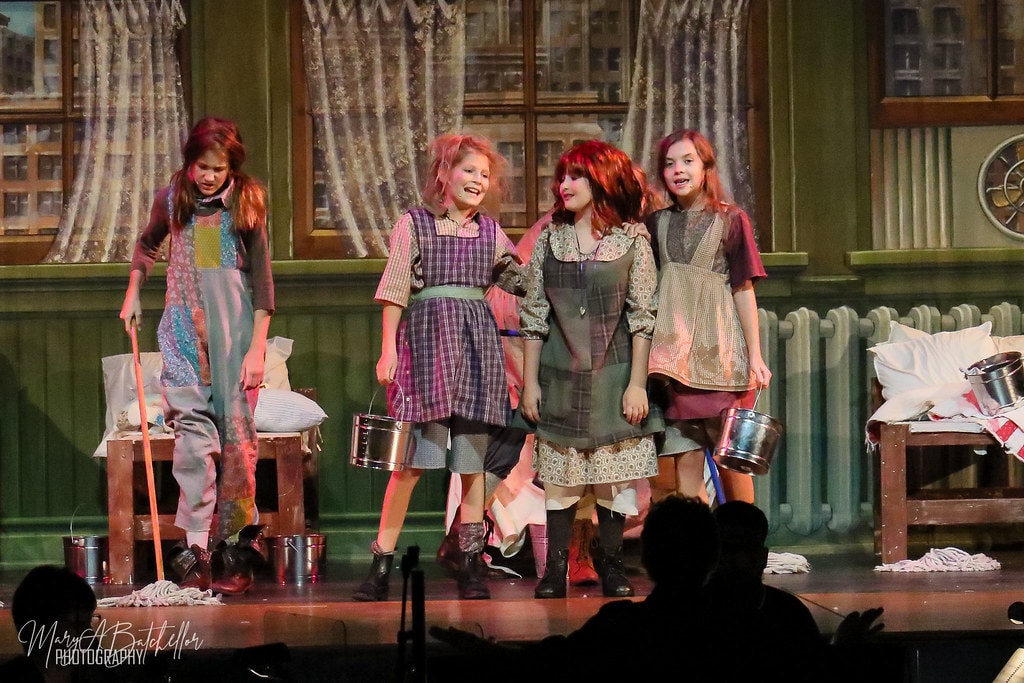 Annie — four orphan girls sing together holding mops and buckets in the orphanage dormitory
