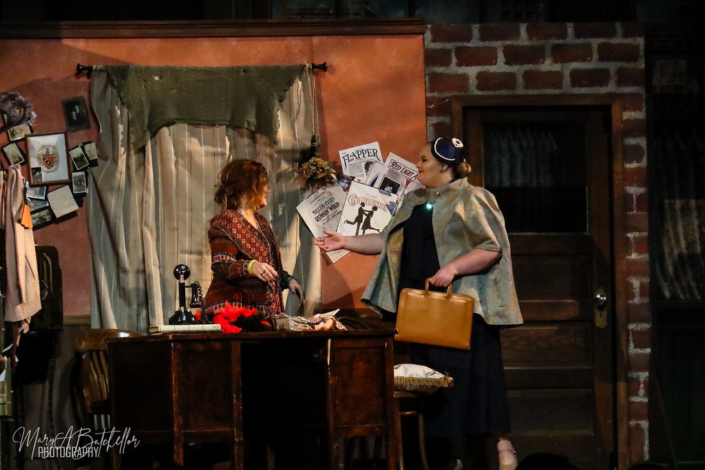 Annie — Miss Hannigan faces a woman in a navy cape and pillbox hat at the orphanage office desk