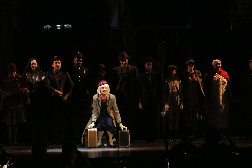 Annie — Grace Farrell kneels center stage with two suitcases — wider angle showing the complete cast
