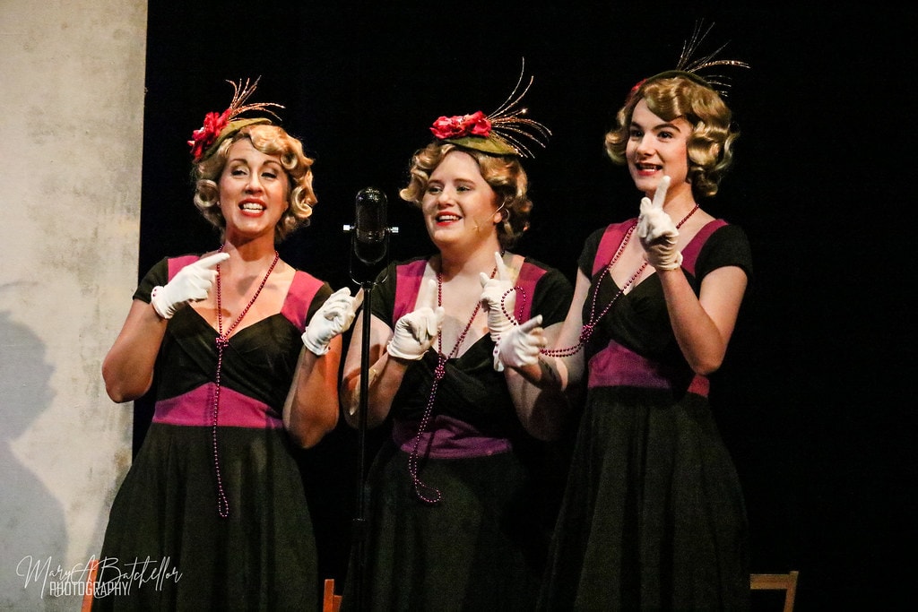 Annie — three women in matching black and magenta 1930s dresses with feathered hats sing a trio