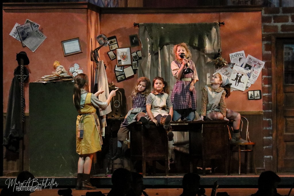 Annie — four orphan girls on a wooden cabinet as a fifth girl points at a vintage radio