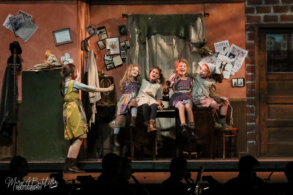 Annie — five orphan girls laugh and sing together on a wooden cabinet