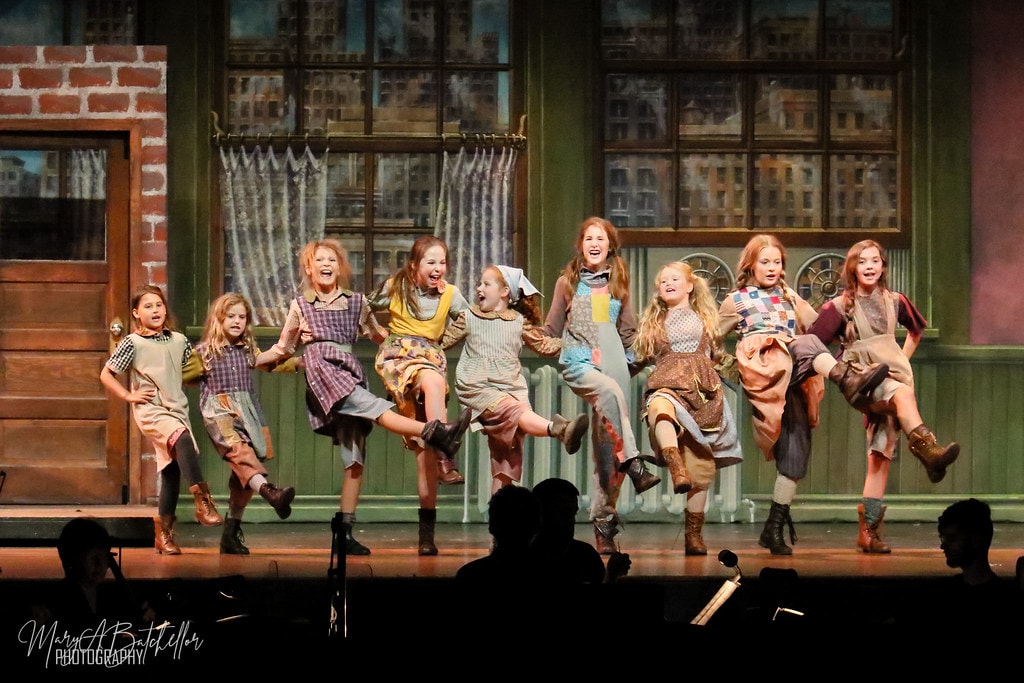 Annie — nine orphan girls hold hands and kick their legs in a joyful chorus line
