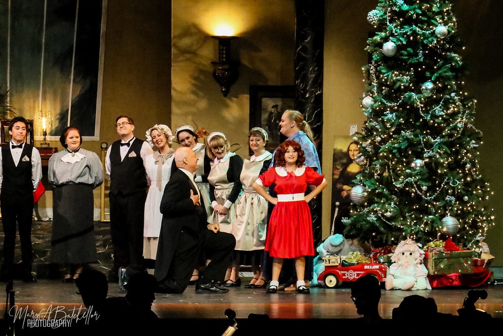 Annie — Oliver Warbucks kneels before Annie in her red Christmas dress as the mansion staff stands behind them
