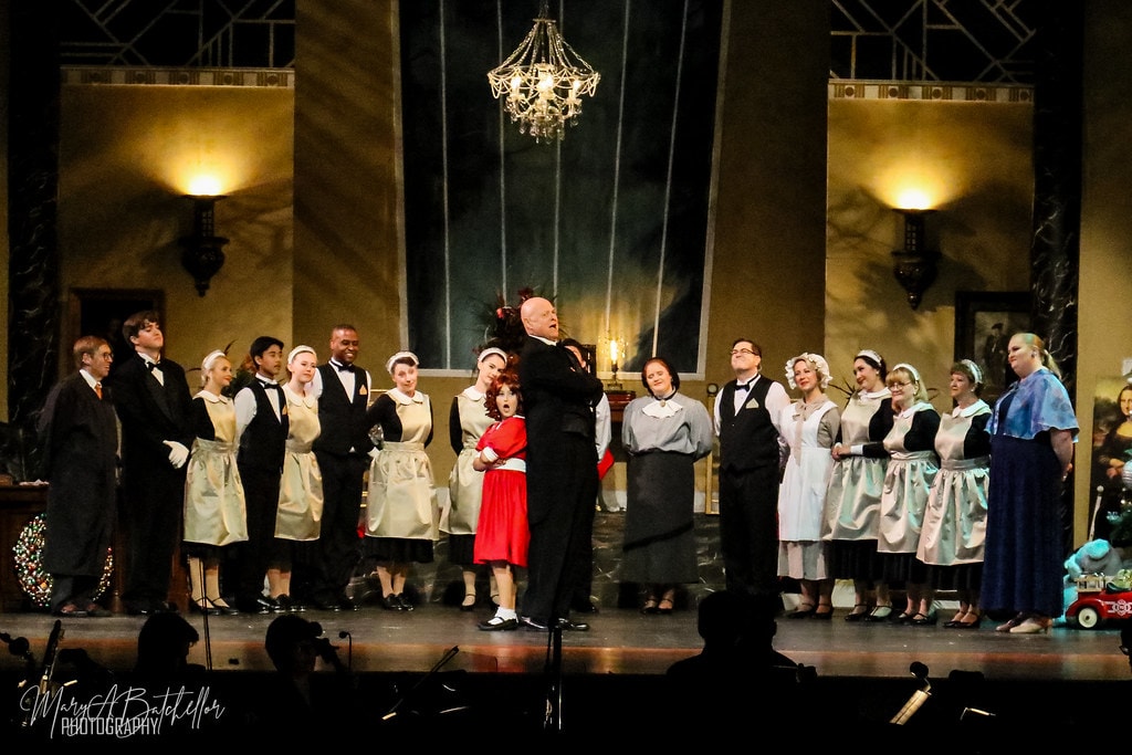 Annie — Oliver Warbucks belts a solo center stage as Annie stands beside him under the chandelier