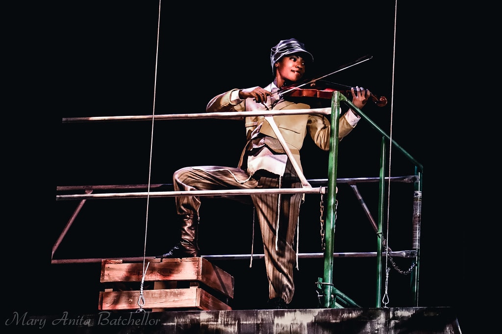 Fiddler on the Roof — the Fiddler in a tan suit and grey cap plays violin on the scaffolding rooftop set against a black backdrop