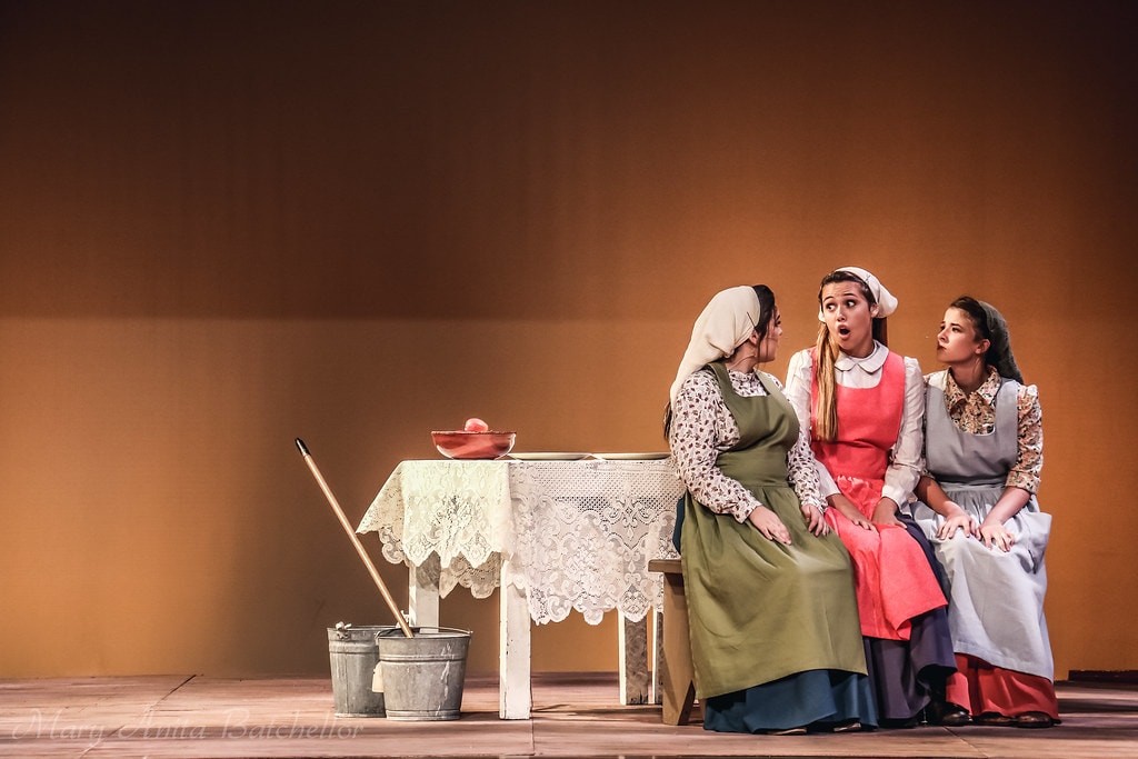 Fiddler on the Roof — three daughters sit at the lace-covered kitchen table in a hushed, wide-eyed conversation scene