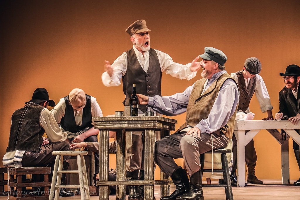 Fiddler on the Roof — two men argue animatedly at a tavern table surrounded by other villagers in the lively inn scene