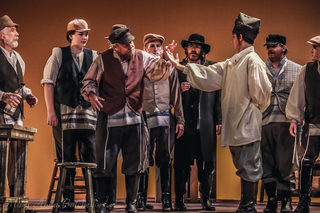 Fiddler on the Roof — a young man in a white shirt gestures firmly at Tevye and a group of male villagers in period caps and vests before the warm amber backdrop