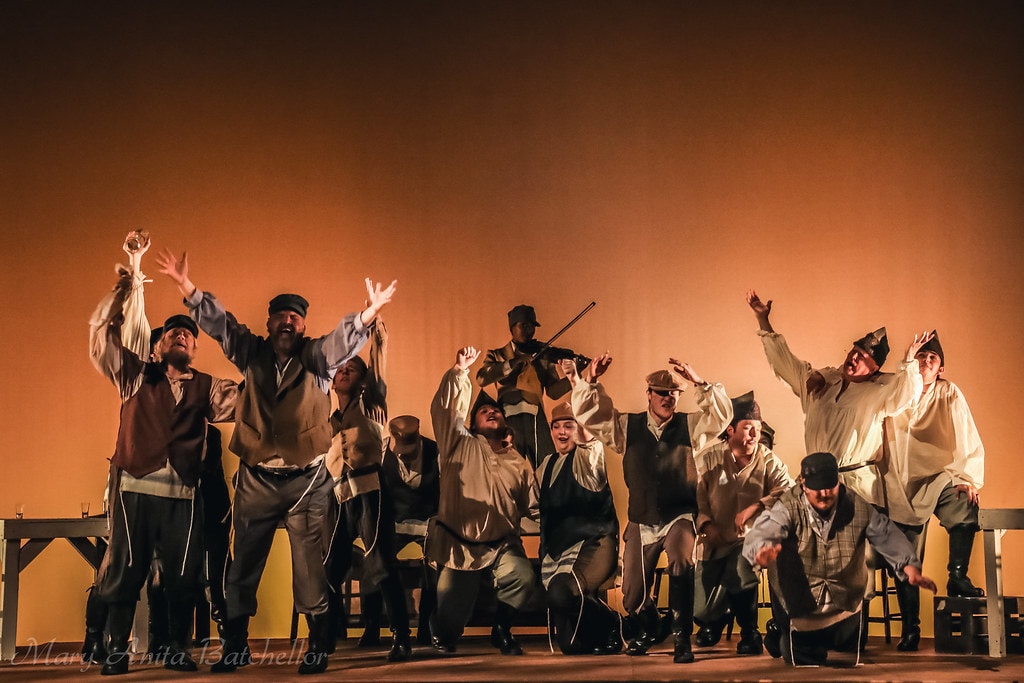 Fiddler on the Roof — the full male ensemble with arms raised joyfully, the Fiddler silhouetted behind them playing violin against the warm amber backdrop