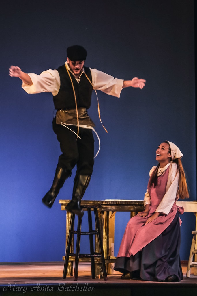 Fiddler on the Roof — a young man in a black vest leaps exuberantly off a wooden stool while a young woman in a pink dress and apron watches and laughs against a deep blue backdrop