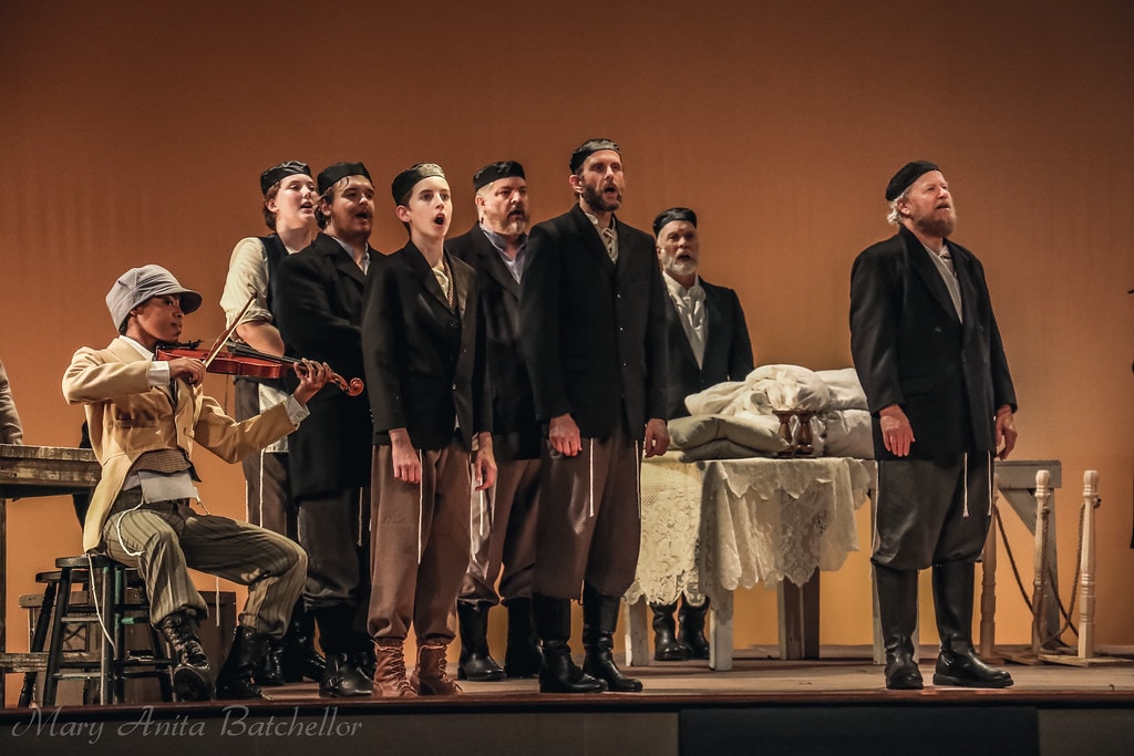 Fiddler on the Roof — the Fiddler plays violin at left while seven men in dark coats and caps sing together beside a lace-covered table against the warm amber backdrop