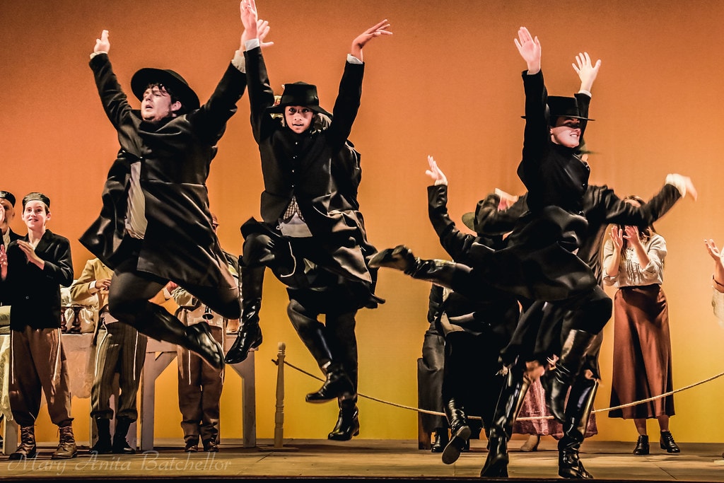 Fiddler on the Roof — three men in black coats and wide-brimmed hats leap exuberantly into the air during the energetic wedding celebration dance