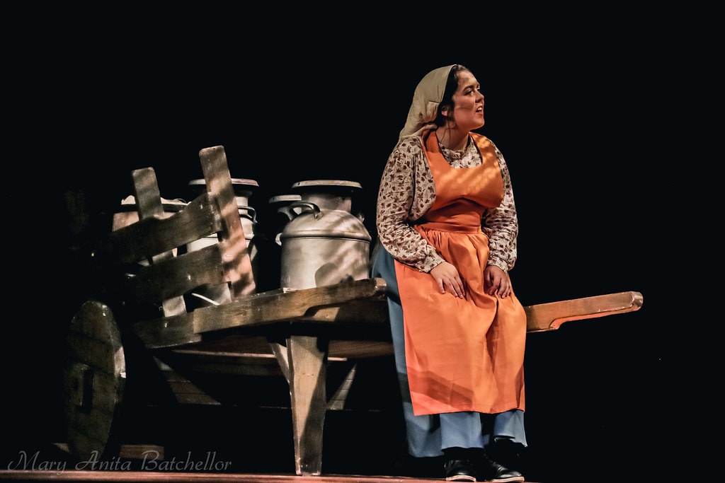 Fiddler on the Roof — Golde in an orange apron and floral blouse sits on the wooden milk cart beside the milk cans, singing a solo in a warm spotlight against a black backdrop