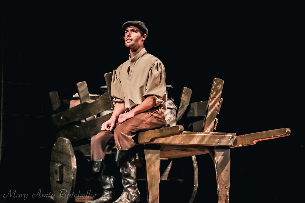 Fiddler on the Roof — a young man in a tan peasant shirt, grey cap, and tall boots sits on the wooden milk cart singing a heartfelt solo against a black backdrop