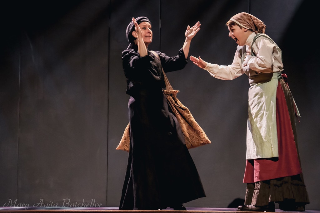 Fiddler on the Roof — Yente the Matchmaker in a black coat gestures dramatically with raised hands toward a young woman in a green apron and red skirt in a lively argument scene