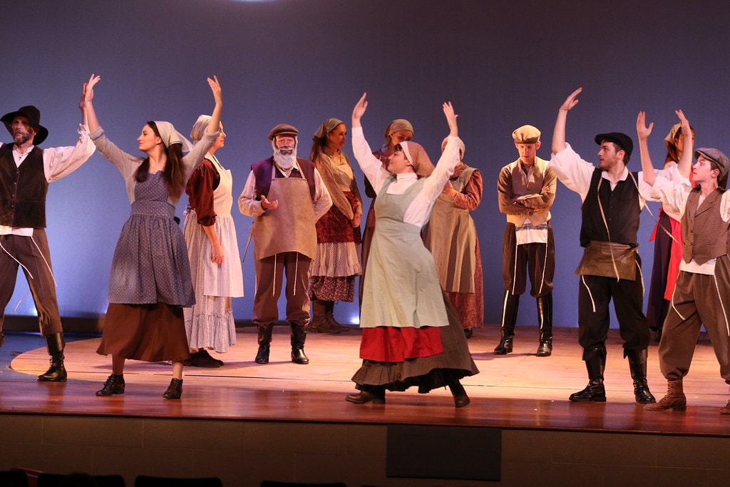 Fiddler on the Roof — the full mixed ensemble raises their arms skyward in a lively dance number on the warm wooden stage floor against a blue backdrop