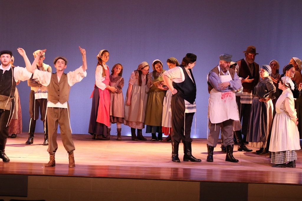 Fiddler on the Roof — a young man gestures upward with one arm as the full ensemble of villagers in period costumes gathers across the warm wooden stage