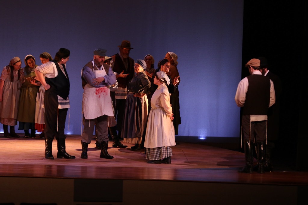 Fiddler on the Roof — a group of villagers including the Butcher in a bloodied apron and Yente in a white bonnet converse in a cluster while a young man stands apart at right against the blue backdrop