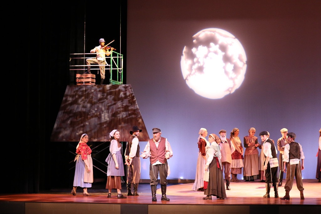Fiddler on the Roof — the Fiddler plays violin atop the rooftop scaffolding as Tevye addresses the full ensemble gathered below before the large glowing moon backdrop