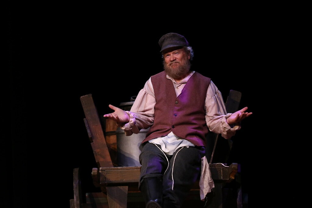 Fiddler on the Roof — Tevye in a burgundy vest and grey cap sits on his wooden milk cart with hands spread wide in a warm close portrait against a black backdrop