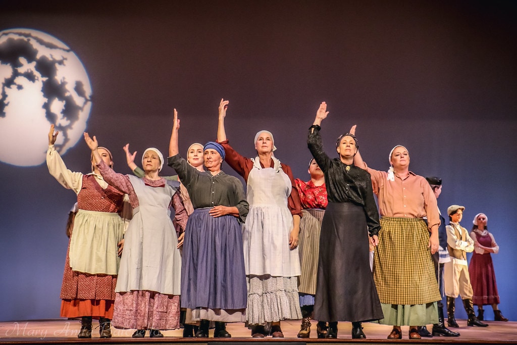 Fiddler on the Roof — the full female ensemble in headscarves and aprons raises their arms before the moon backdrop