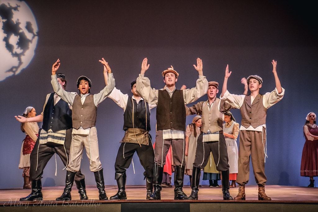 Fiddler on the Roof — five young men in vests and caps raise their arms and sing together before the moon backdrop