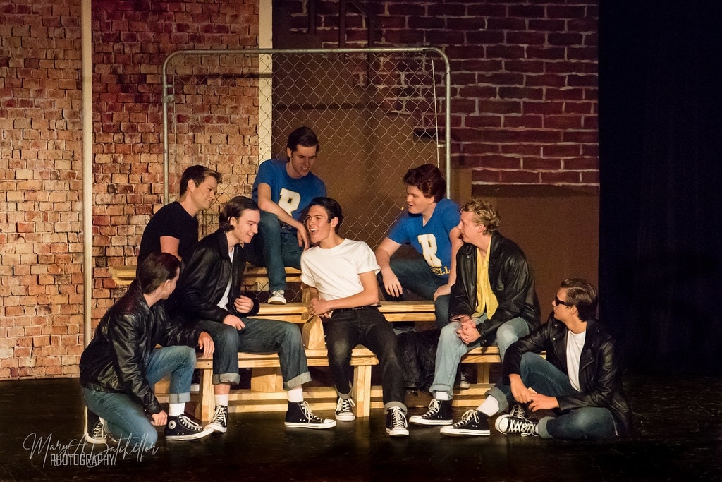 Grease — T-Birds lounging on bleachers by chain-link fence