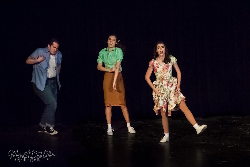Grease — three performers dancing and singing on dark stage
