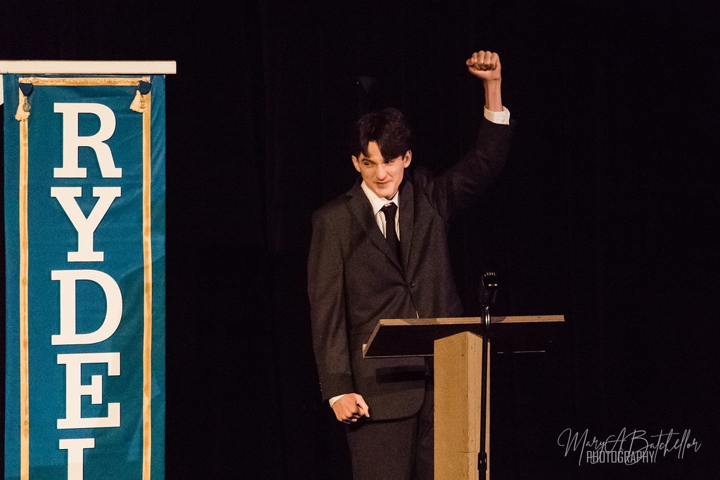 Grease — Danny Zuko at podium raising fist by Rydell banner