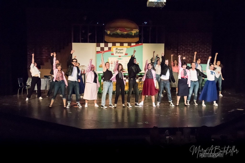 Grease — Danny at Rydell podium raising fist with banner close-up