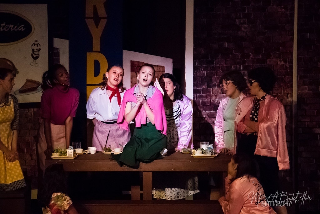 Grease — Sandy singing on cafeteria table surrounded by Pink Ladies