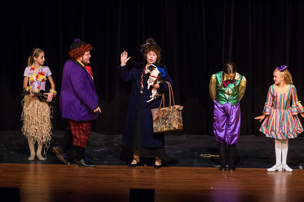 Gypsy — five performers on the bare black stage: a girl in a Hawaiian hula skirt with ukulele, the boy in the purple plaid suit, Momma Rose in a navy coat holding a small dog and a tapestry bag with one hand raised, a child bowing in a green-and-gold costume, and a girl in a rainbow-striped dress