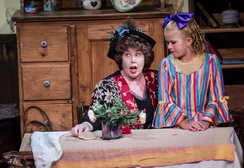 Gypsy — Momma Rose in a black feathered hat and floral-print jacket sits at a kitchen table with a vase of roses, her mouth open in shock, as a young girl in a rainbow-striped dress with a large purple bow stands beside her in the realistic kitchen set