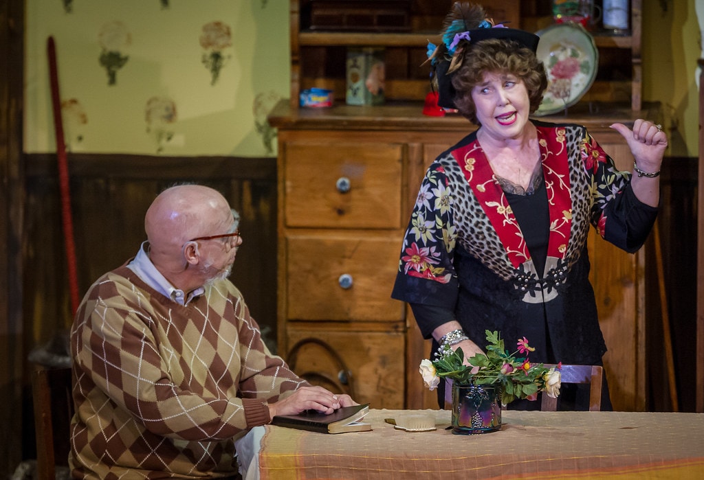 Gypsy — Momma Rose in her black feathered hat and floral-print jacket stands at the kitchen table gesturing with a thumbs-up as a bald man in a brown argyle sweater sits reading a book in the realistic kitchen set