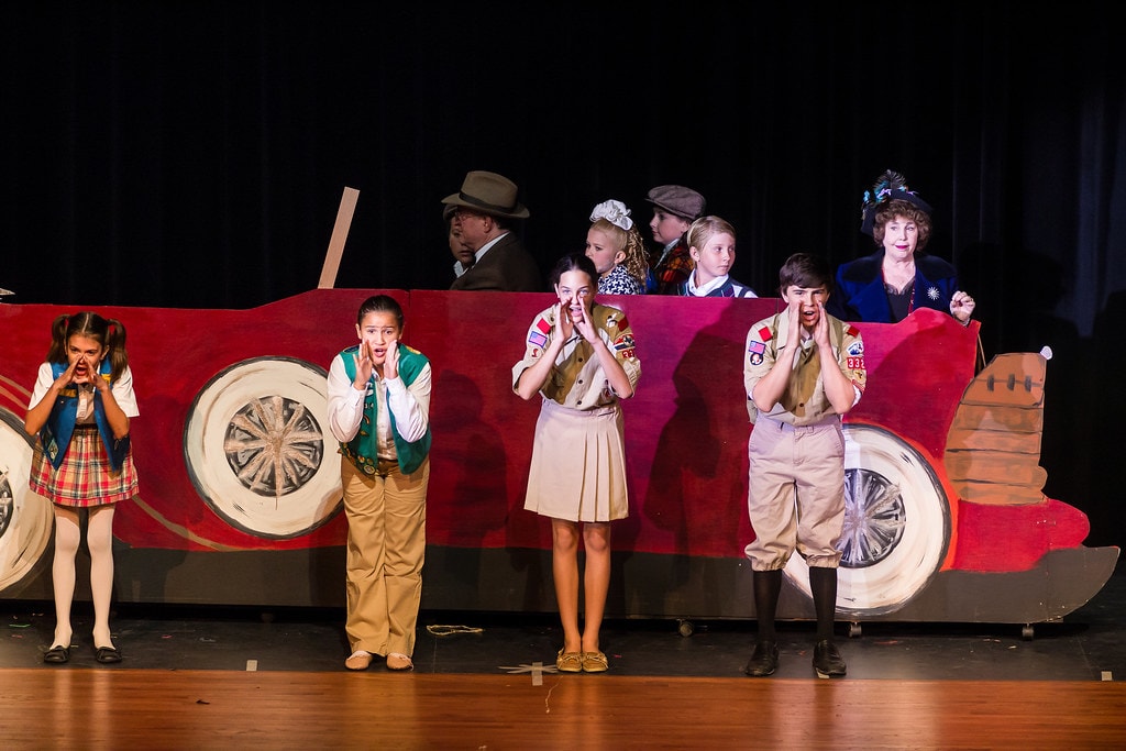 Gypsy — four children in scout uniforms and a girl in a plaid dress stand in front of a large red vintage car prop with hands on cheeks in exaggerated shock, while Momma Rose in a navy coat and two other performers ride in the car above them