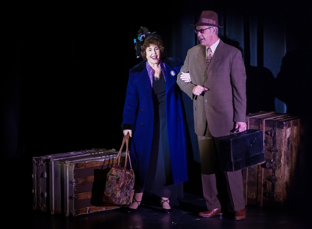 Gypsy — Momma Rose in her navy coat with a peacock-feathered hat laughs warmly as she walks arm-in-arm with a man in a brown suit and hat carrying a vintage suitcase, both lit by a dramatic blue spotlight against a dark stage