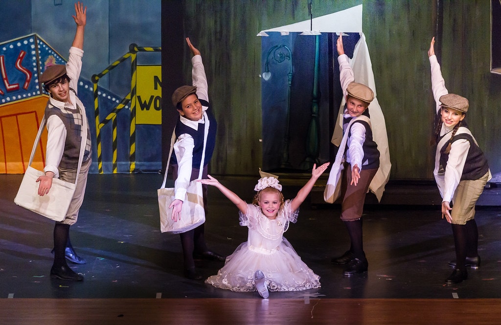 Gypsy — five children in newsboy caps and grey vests with canvas bags pose in a dynamic dance formation before a colorful Broadway cityscape backdrop with neon signs, a small girl in a white lace dress at center with arms raised wide