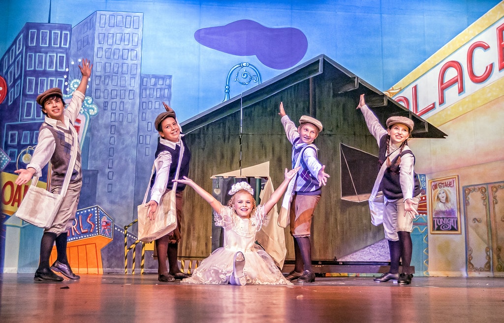 Gypsy — the same five newsboy children in a wider shot before the full Broadway Palace backdrop with skyscrapers and marquee signs, the girl in the white lace dress at center with arms spread joyfully