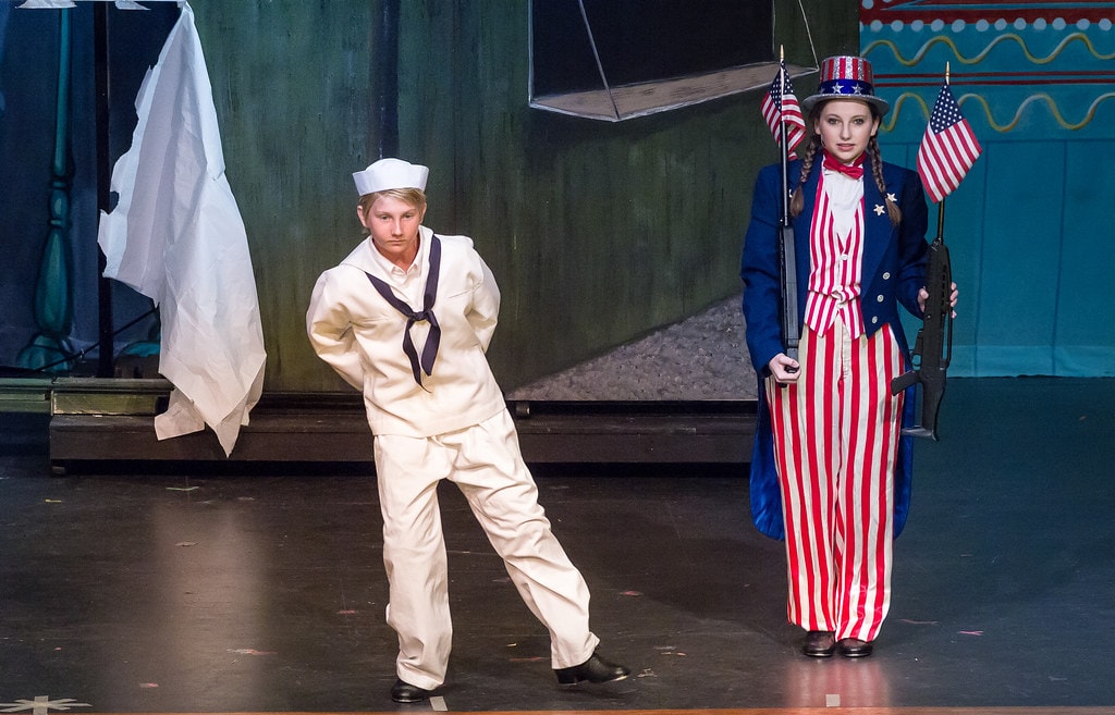 Gypsy — a teenage boy in a white sailor uniform leans forward with hands on hips before a teal circus backdrop, while a teenage girl in a patriotic red-white-and-blue striped Uncle Sam costume with top hat holds two American flags and a rifle