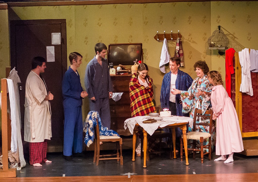 Gypsy — six performers in pajamas and robes gather around a table with a birthday cake in the realistic boarding house set with green floral wallpaper, a dresser with a clock, and a birdcage on the wall