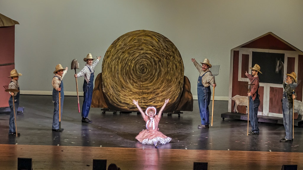 Gypsy — a girl in a pink gingham dress and bonnet kneels center stage with arms raised joyfully before a giant round hay bale prop as six men in denim overalls and straw hats hold shovels and rakes in the farm vaudeville act